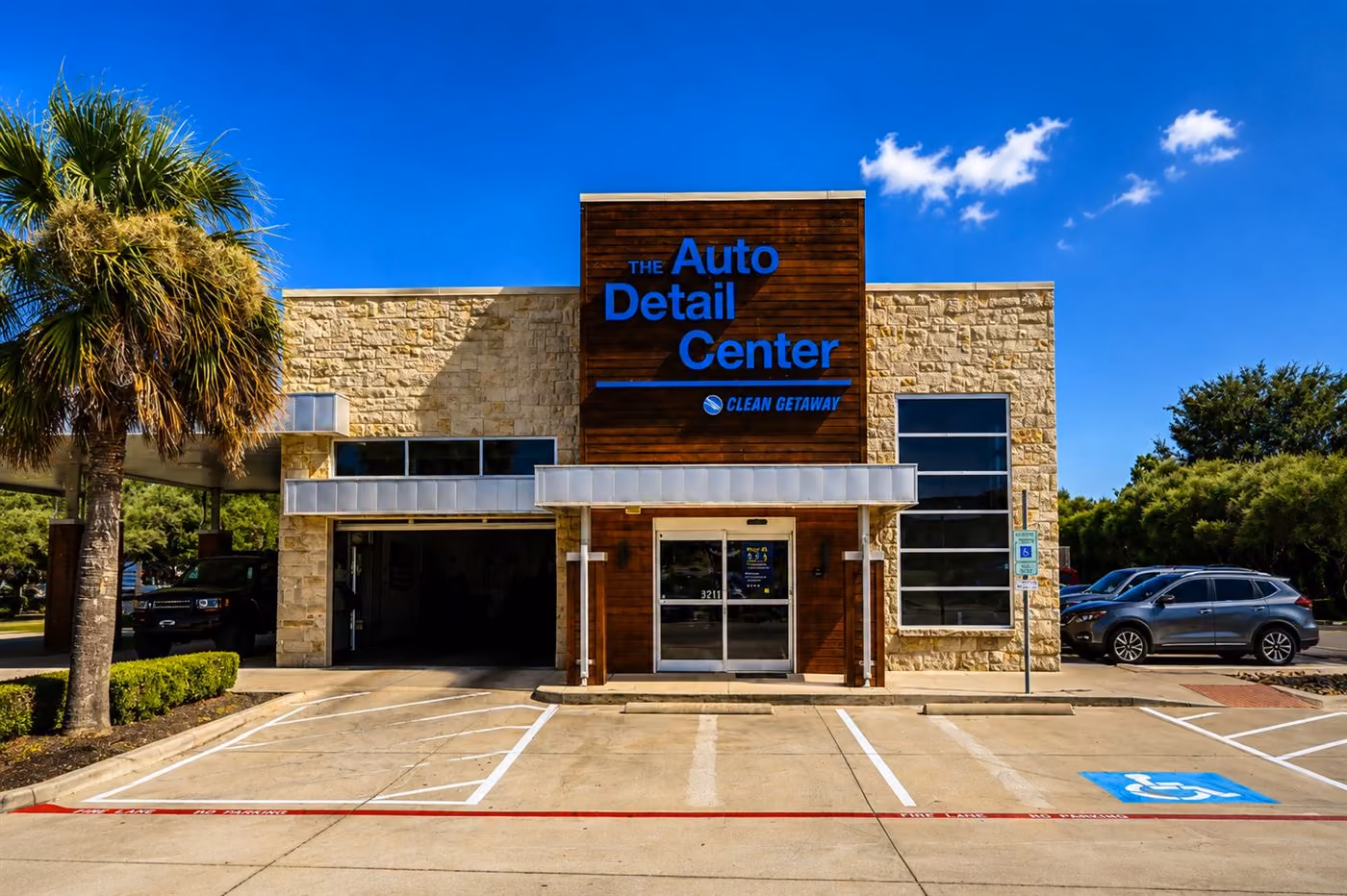 The Auto Detail Center building — stone and wood facade with blue signage, open bay door, and palm trees under a blue sky