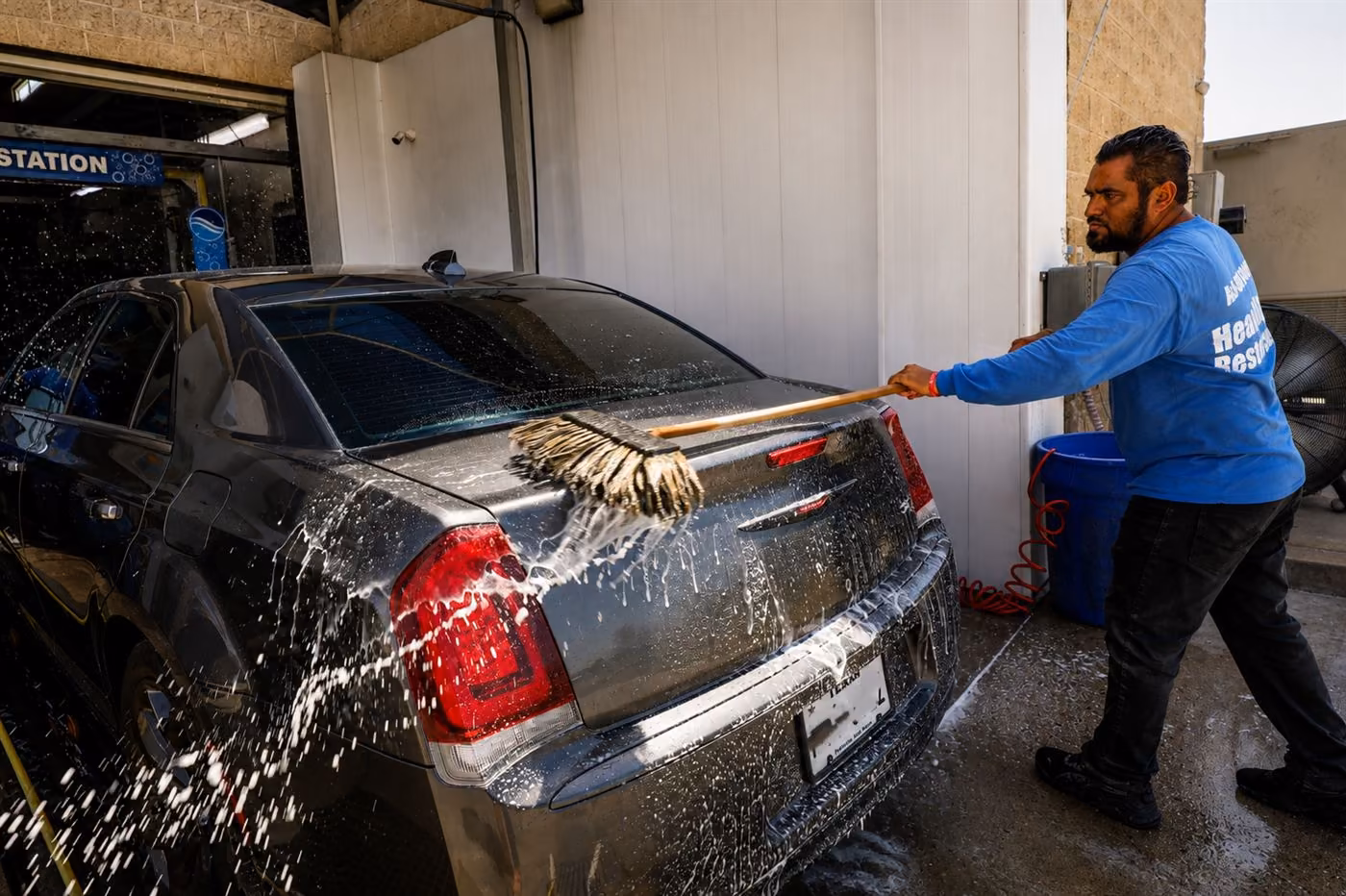 Crew member hand-washing the rear of a dark sedan with a soft mitt as water sprays